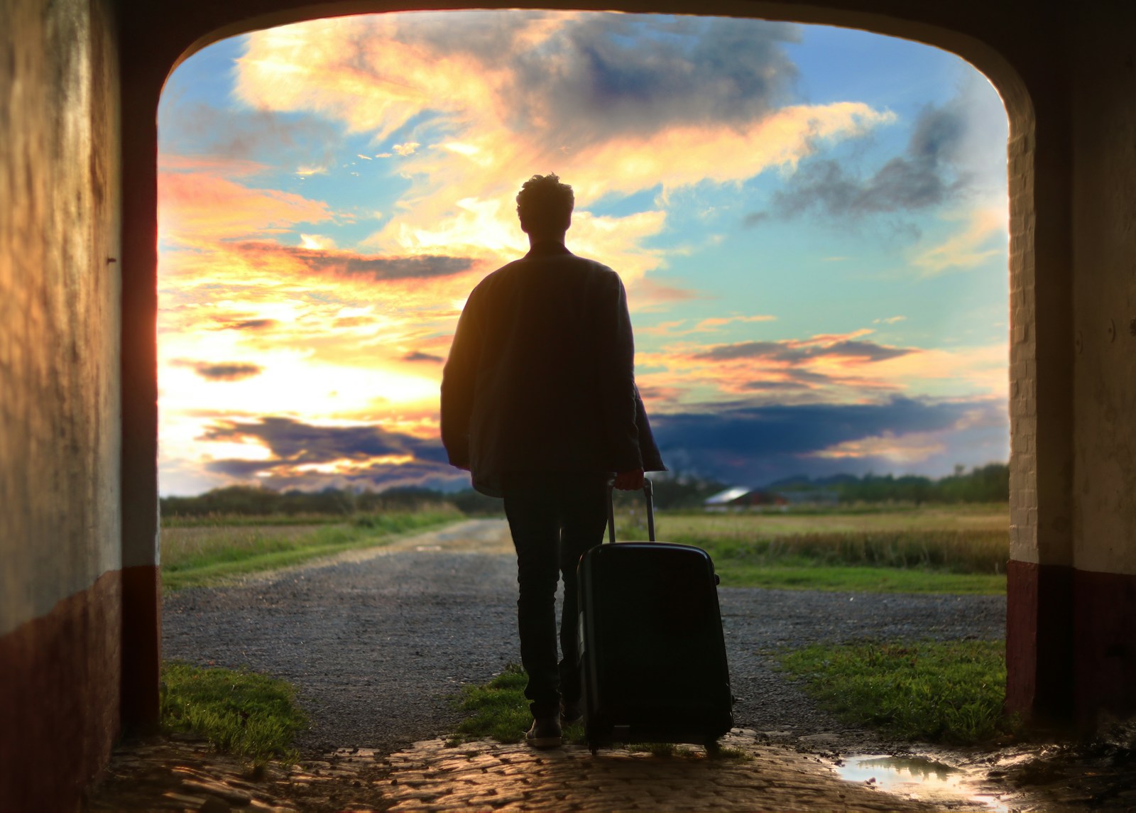 Person with suitcase walking towards a vibrant sunset through a brick archway, symbolising travel and new beginnings