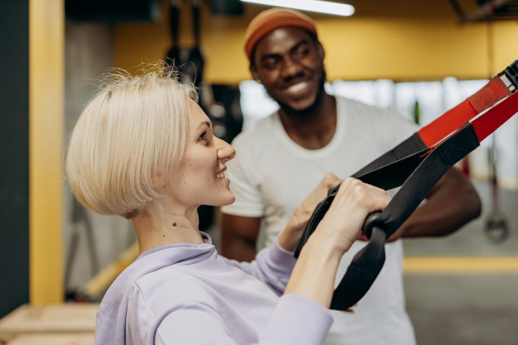 pexels-photo-6740311-6740311 A smiling fitness trainer assists a woman during a TRX workout session indoors, promoting a healthy lifestyle.