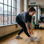 Woman stretching in a gym next to large windows, with exercise equipment and people in the background