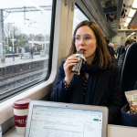 Woman drinking on a train, laptop open, snacks in bag, approaching Woking station on a rainy day